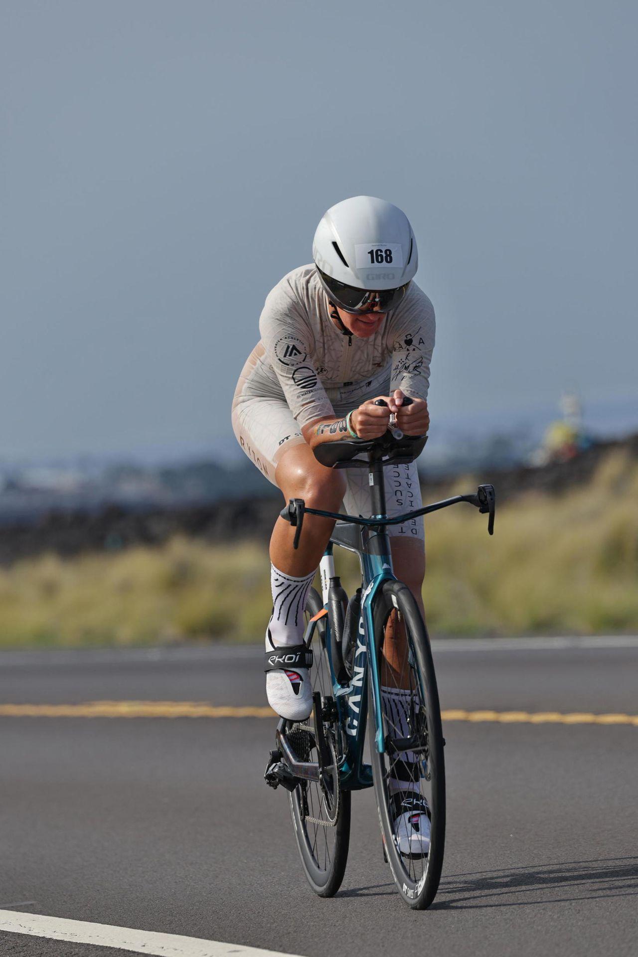 Cyclist in aerodynamic gear and helmet riding a racing bicycle on an empty road surrounded by grass.